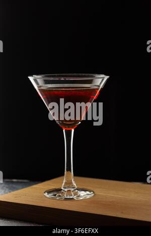 A classic martini glass filled with a deep red cocktail stands on a wooden board against a black backdrop, with dramatic red light reflecting along th Stock Photo