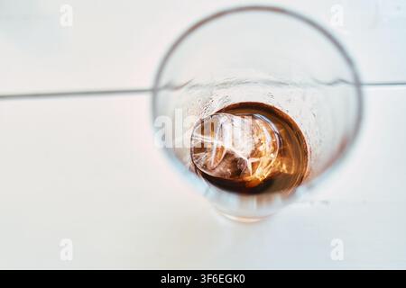 High angle close-up shot of a glass containing cola and a single large melting ice cube on a white wooden table surface. Stock Photo