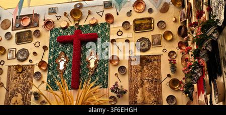 Red floral cross displayed on a decorated wall with copper cookware, ceramic plates, framed artwork and patterned textiles as part of a festival insta Stock Photo