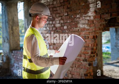 Engineer in safety gear reviewing blueprints on construction site. Architectural inspection project planning and renovation work. Stock Photo