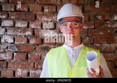 Architect holding blueprints against brick wall at a construction site. Professional portrait of an architect in the construction industry. Stock Photo