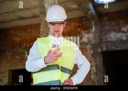 Construction engineer wearing safety gear checks updates on smartphone at a building site. Mobile technology and site progress monitoring in construct Stock Photo