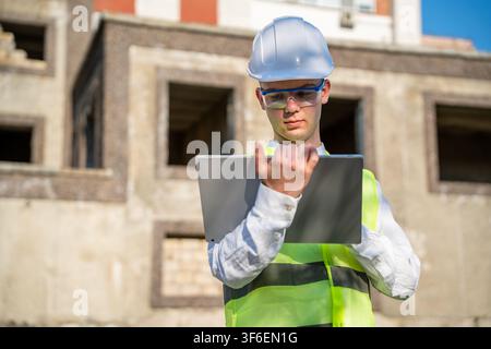 Engineer using laptop on construction site wearing safety helmet and vest. Mobile technology and modern construction business solutions. Stock Photo