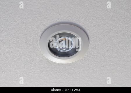 A close-up view of a circular recessed light with a bright LED bulb, installed in a textured white ceiling Stock Photo