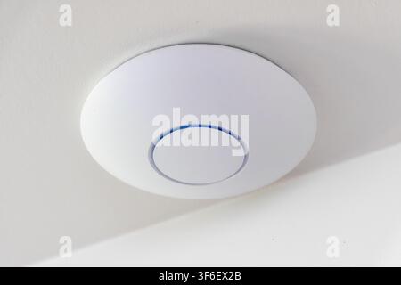 A sleek, circular Wi-Fi device with a subtle blue LED indicator is discreetly installed on a white ceiling surface Stock Photo