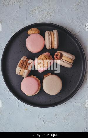 Selection of colorful French macarons decorated with cookies, mini chocolate bars, and pretzel on black plate, top view Stock Photo