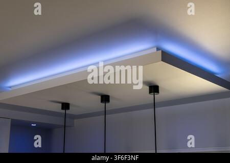 A contemporary interior ceiling features a layered design with subtle blue LED illumination highlighting architectural details and three hanging lamps Stock Photo
