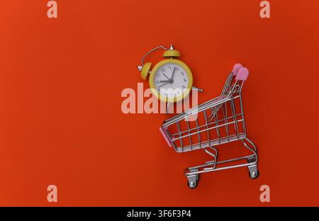 Mini supermarket trolley and retro alarm clock on orange background. Shopping time. Top view. Stock Photo