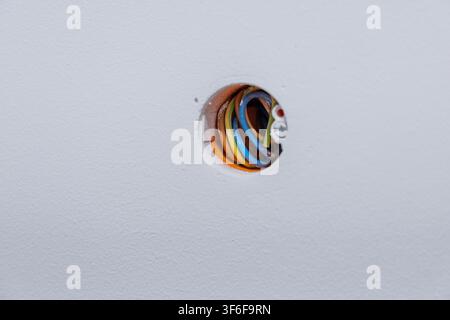 A circular hole in a white textured wall reveals a bundle of multicolored electrical wires, hinting at construction or repair work Stock Photo