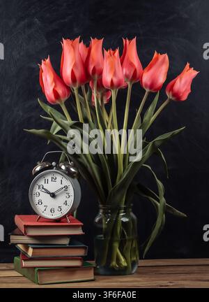 Bouquet of red tulips and stack of books with alarm clock on chalk board background. School still life Stock Photo