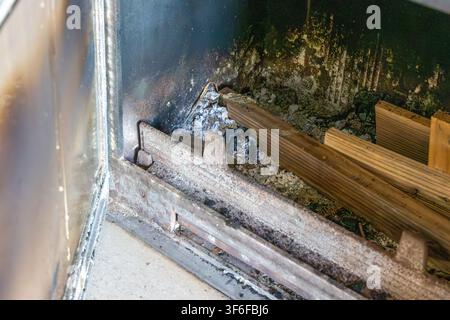 The interior of a fireplace shows a metal grate covered in ash and soot, with several pieces of wood ready for burning Stock Photo