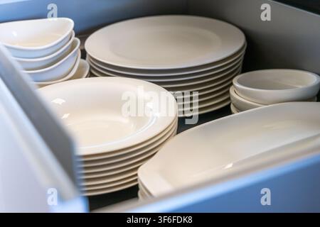 A collection of clean, modern white dishes including dinner plates, soup bowls, and serving dishes are stored in a tidy kitchen cabinet Stock Photo