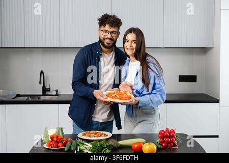 Young smiling couple preparing a homemade pizza with fresh vegetables in a modern kitchen, sharing a healthy meal and togetherness Stock Photo