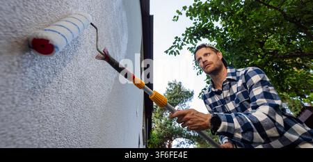 House painter applying protective coating on house exterior wall with roller. Facade protection against algae, moss and fungal Stock Photo