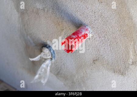 A red plastic conduit fitting is inserted into a textured concrete wall, with a blue ring and plastic debris visible nearby Stock Photo