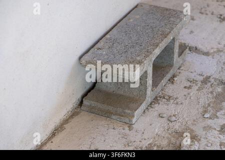 A single cinder block is placed against a rough, off-white wall on a gritty, concrete surface with scattered debris Stock Photo