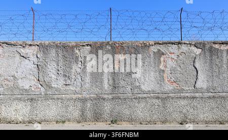 Partially peeled rough concrete wall with some bricks exposed and rusty barbed wire on top. Blue sky above. Grunge background for copy space. Stock Photo