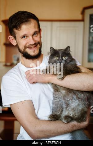 Happy bearded man cradles his fluffy grey cat in an indoor setting, highlighting the bond of friendship and companionship between pet and owner Stock Photo