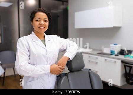 Confident black female dentist posing beside dental chair in modern clinic Stock Photo