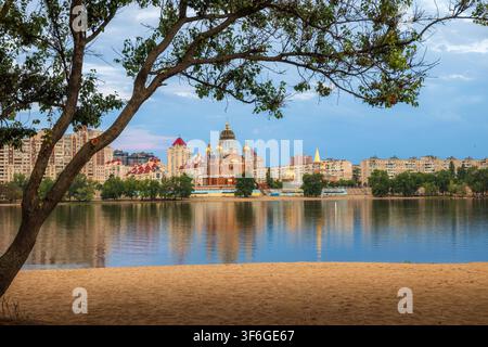 Scenic view of Obolon Embankment in Kyiv, Ukraine, after a summer thunderstorm before sunset, with calm reflections, sandy beach, leafy tree framing, Stock Photo
