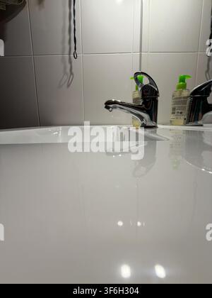 Chrome mixer tap and white bathroom basin reflected in a clean glossy surface against tiled wall Stock Photo