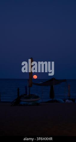 Red Moon Rising Over a Quiet Seaside Beach at Night with Closed Sun Umbrellas Stock Photo