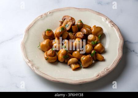 Sautéed whole button mushrooms with garlic and fresh herbs served on a vintage ceramic plate over white marble background Stock Photo