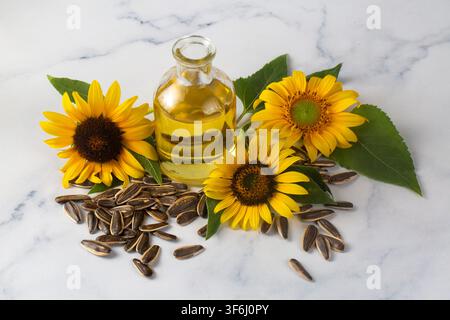 Clear glass bottle of golden sunflower oil with fresh yellow flowers and black seeds on white marble background Stock Photo