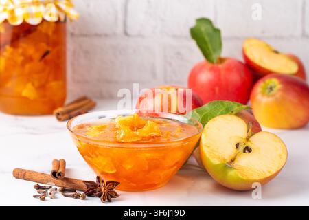 Golden apple jam in a glass bowl with fresh red apples and cinnamon sticks on white marble background Stock Photo