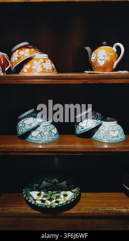 Tashkent, Uzbekistan - April 18, 2026 - Display of orange and turquoise ceramic teapots and bowls with floral designs on dark wood shelves perfect for Stock Photo