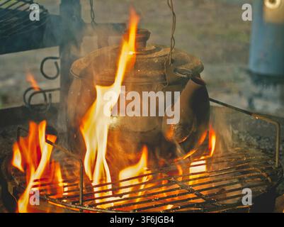 Paimela, Finland - 17.06.2017: Weathered black iron teapot heating on a metal grate above intense campfire flames in a rural setting Stock Photo