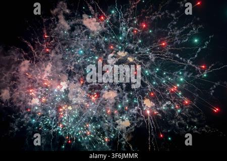Colorful fireworks exploding in the night sky with vibrant red and teal (cyan) sparks, creating smoke trails during a pyrotechnic celebration display. Stock Photo