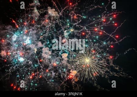 A spectacular pyrotechnic display in the night sky featuring a bright golden starburst on the lower right, surrounded by red and cyan sparks and glowi Stock Photo