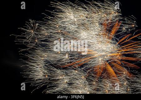Abstract and dramatic pyrotechnic explosion in the night sky featuring dense white and golden sparkling trails layered over glowing orange firework ra Stock Photo