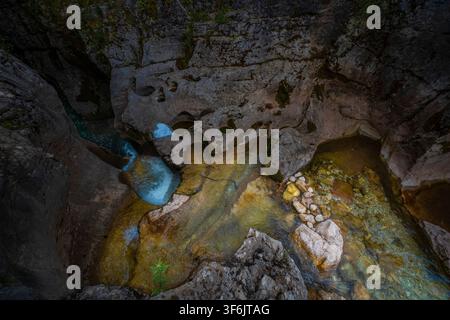 The image showcases a steep, rocky cave with mineral-rich water flowing through, exhibiting vibrant yellow, blue, and green hues amidst textured gray Stock Photo