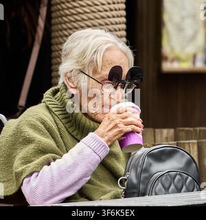 An elderly woman with flip-up sunglasses attached to her spectacles drinks from a paper coffee cup outdoors Stock Photo