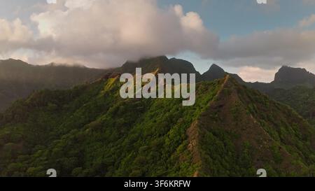 Lush green volcanic mountain ridges rise under an epic cloudy sky, showcasing the dramatic tropical landscape and natural beauty of Moorea in French Polynesia at sunset. Aerial view drone flight Stock Photo