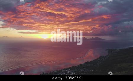 Moorea coastline at dusk, vivid pink, orange and purple sky reflected on calm Pacific waters with silhouetted mountains and tranquil tropical beach. Natural landscape. Aerial view drone flight Stock Photo
