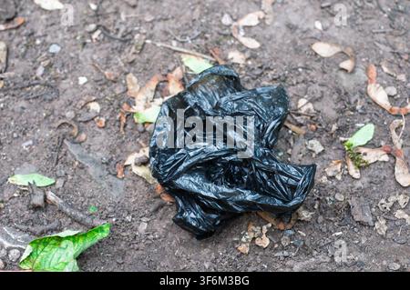 Crumpled black plastic bag on dirt ground. Scattered dry leaves, small stones, and green foliage are visible. Outdoor. Stock Photo