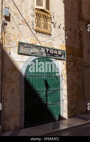 A weathered green "Store" sign hangs above an arched, dark green wooden door beneath an ornate window grille in Valletta, Malta Stock Photo