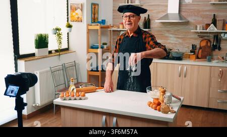 Male baker records a baking demonstration for online viewers beside the kitchen worktop while camera equipment stands beside the kitchen worktop. Stock Photo