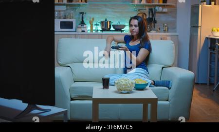At home, the woman changes television channels with a remote control while the television screen stays in view ahead of the sofa during program selection. Stock Photo