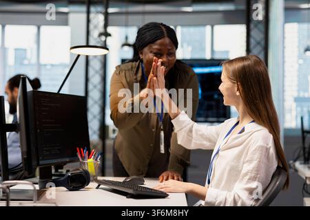 Smiling system administrator mentoring junior dev at desk in office, congratulating her for optimizing code. Joyous tech startup IT specialist doing high five with female developer intern Stock Photo