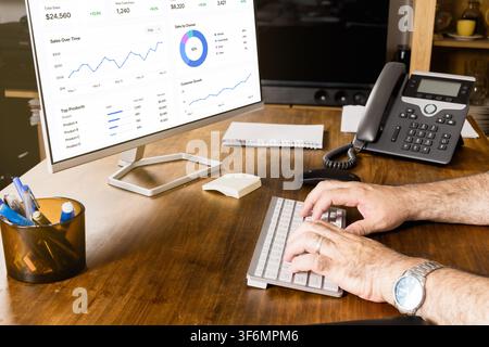 Close-up of a professional typing on a keyboard beside a monitor showing sales charts, customer growth and business analytics on a wooden office desk. Stock Photo