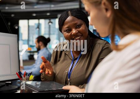 Pleased team leader mentoring junior dev at desk in office, congratulating her for finishing tasks. Satisfied african american IT specialist offering positive feedback to woman doing internship Stock Photo