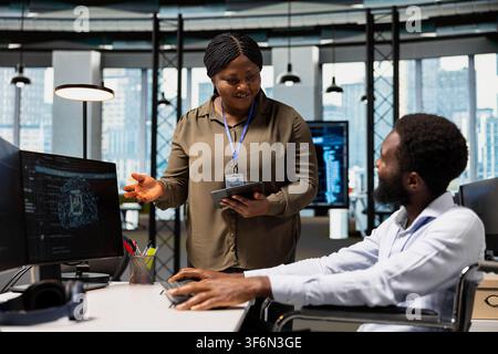 Smiling manager oversees software developer in wheelchair using AI automation, refining algorithms. Jolly supervisor pleased with work done by programmer with paraplegia using artificial intelligence Stock Photo