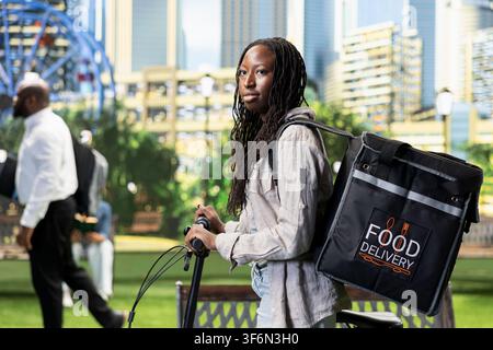 Woman bicycle courier standing with bike and insulated delivery backpack in city park, ready to continue work after checking next route. Immigrant gig worker starting delivery. Stock Photo