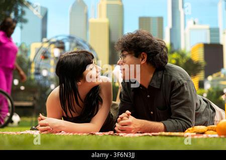 Young couple laying face to face and smiling at each other on picnic blanket, smiling during intimate conversation with food and blurred city skyline. Romantic picnic idea outdoors. Stock Photo