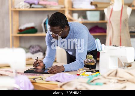 Male tailor draws sketch of future clothing on an electronic tablet. Working with modern gadgets in sewing workshop Stock Photo
