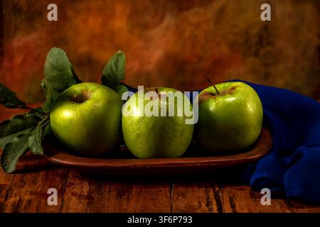 A study of three green apples on a rustic earthenware dish, with a warm background and a splash of blue scarf.. Still life. Stock Photo